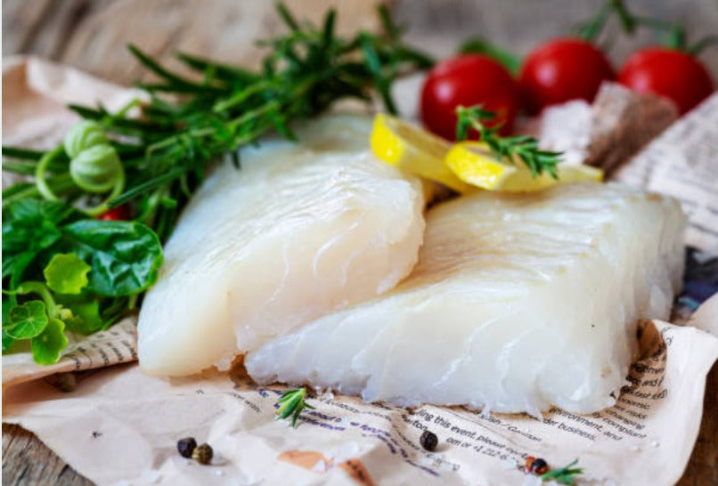 Chilean Sea Bass fillet, 8 oz portion, served with fresh greens, lemon slices, and cherry tomatoes — premium white fish with buttery texture.